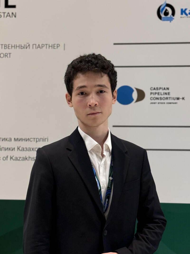 A young man in a black suit and white shirt stands in front of a backdrop featuring logos and text, including "CASPIAN PIPELINE CONSORTIUM-K" and Kazakh text.