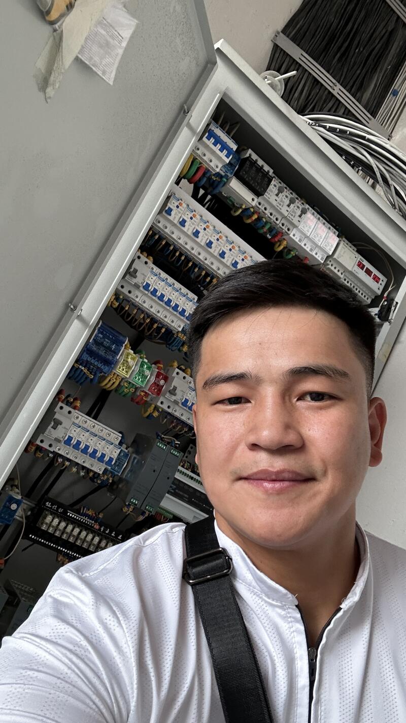 A man in a white shirt with a black strap poses in front of an open electrical panel filled with circuit breakers and wires.