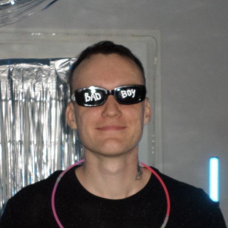 A man wearing black sunglasses with "BAD BOY" written on them smiles at the camera, with a pink glow stick around his neck and a metallic background.