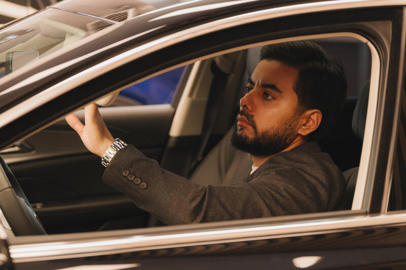 A man with a beard sits in a car, wearing a suit and a watch, while adjusting the rearview mirror.