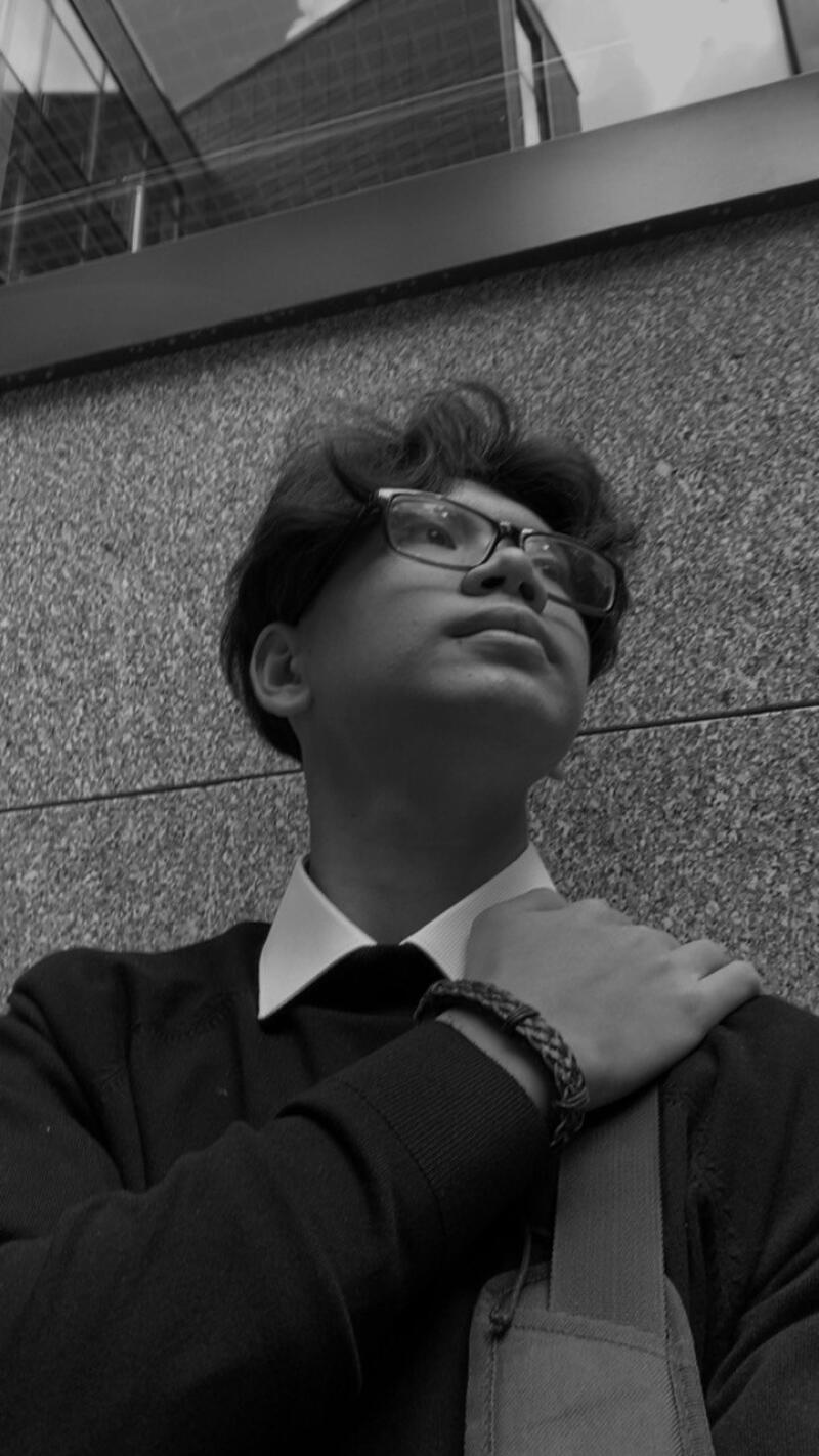 A black and white, low-angle shot of a young person with glasses, wearing a sweater over a collared shirt, standing against a textured wall and looking upwards.