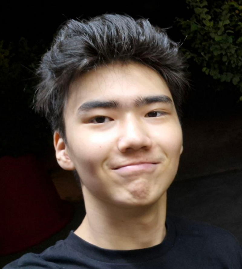 A young man with dark hair and a black shirt is smiling slightly at the camera.