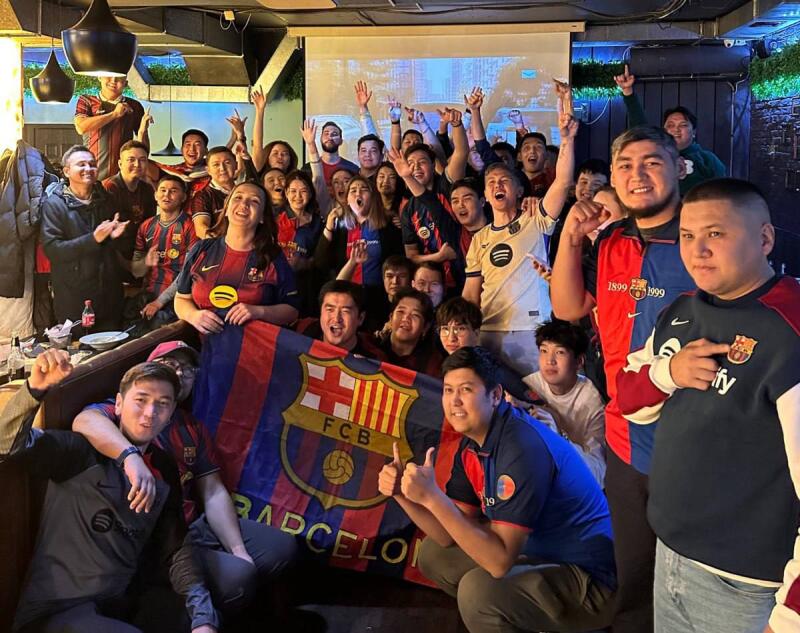 A large group of people are gathered, many wearing FC Barcelona jerseys, and holding a Barcelona flag with the text "BARCELONA" on it.