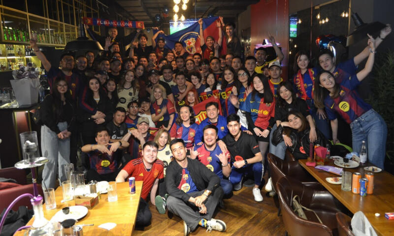 A large group of people are gathered indoors, many wearing FC Barcelona jerseys, with an FC Barcelona banner visible in the background.