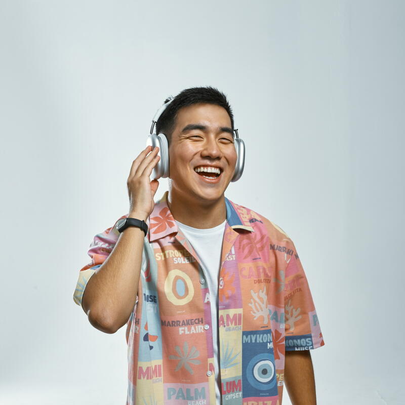 A smiling Asian man wearing headphones and a colorful patterned shirt with words like "STRO SOLEIL", "MARRAKECH", and "MYKONOS" on it, stands against a plain background.