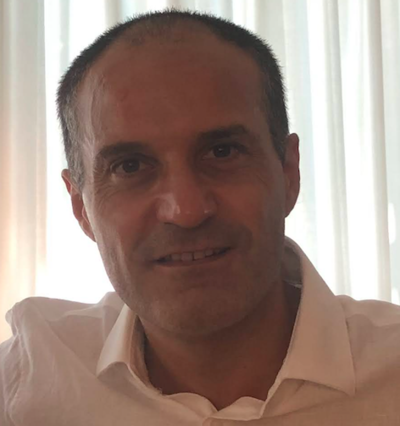 A middle-aged man with short dark hair is wearing a white collared shirt.