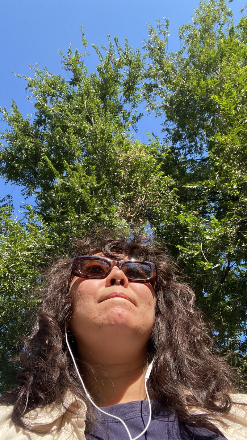 The low-angle shot captures a person wearing sunglasses and earphones, with a backdrop of lush green trees against a clear blue sky.