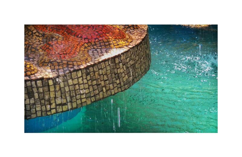 A mosaic fountain with red, yellow, and brown tiles, cascading water, and a turquoise pool.