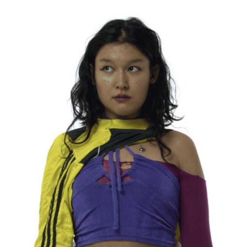 A woman with shoulder-length dark hair looks to the side wearing a purple top with cutouts, a yellow and black jacket, and a magenta sleeve on one arm.