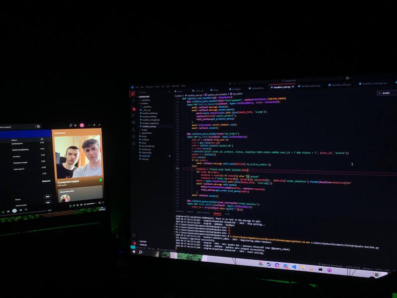 The photo shows two computer screens in a dark room: the left screen displays a music streaming application with a photo of two young men, while the right screen shows lines of code.
