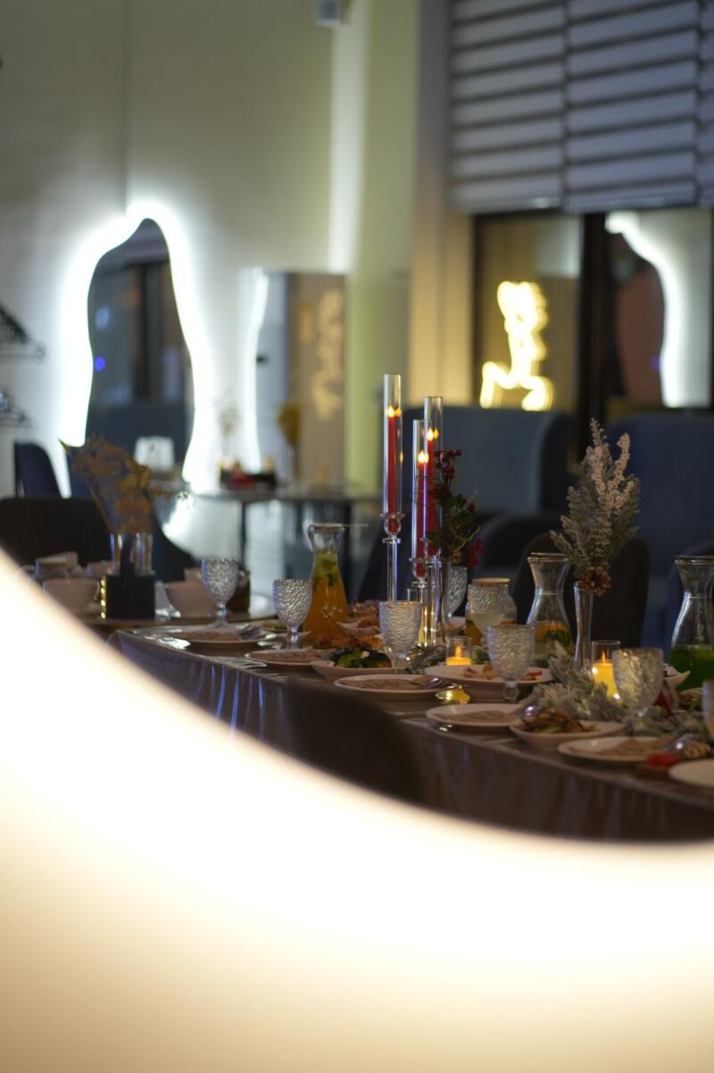 A long table is set for a meal with plates, glasses, candles, and carafes of juice, with decorative elements, under soft lighting with a blurred foreground.