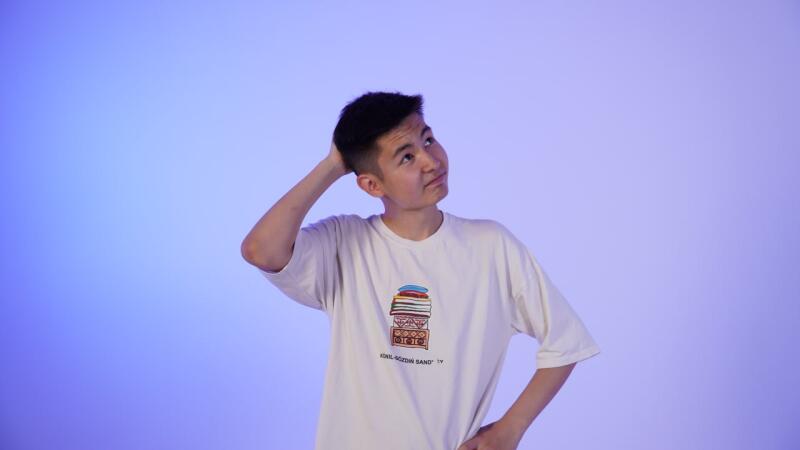 A young man with short dark hair is looking up thoughtfully while scratching his head, wearing a white t-shirt with a graphic design.