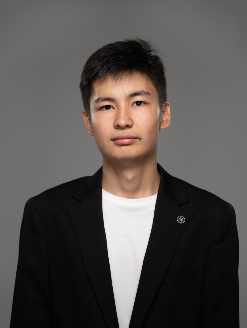 A portrait of a young man with short black hair, wearing a white t-shirt and a black suit jacket, against a grey background.