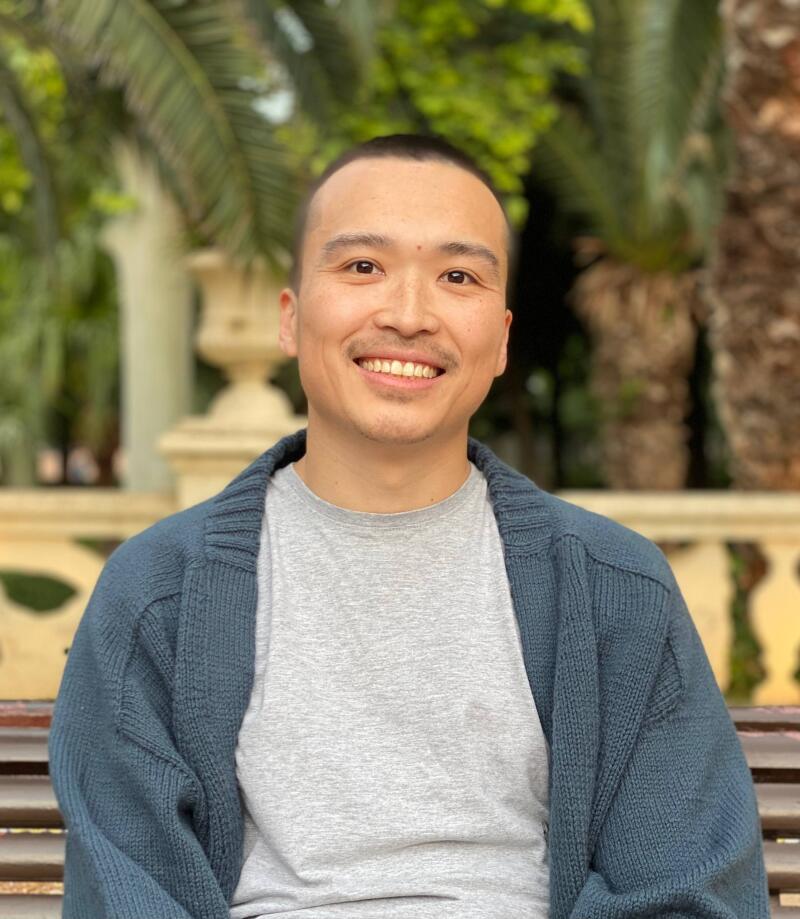 A smiling Asian man is sitting on a bench, wearing a gray T-shirt and a dark blue cardigan, with palm trees in the background.