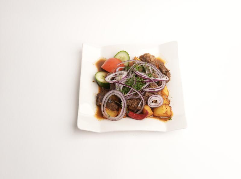 На белой тарелке блюдо с мясом, картофелем, огурцами, помидорами и кольцами красного лука, приправленное укропом и красным перцем чили.
