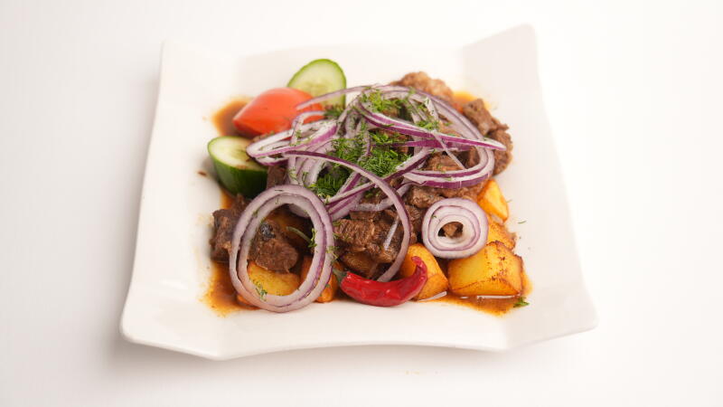 На белой тарелке блюдо из мяса и картофеля, сверху украшенное кольцами красного лука и укропом, в сопровождении нарезанного огурца, помидора и красного перца чили.