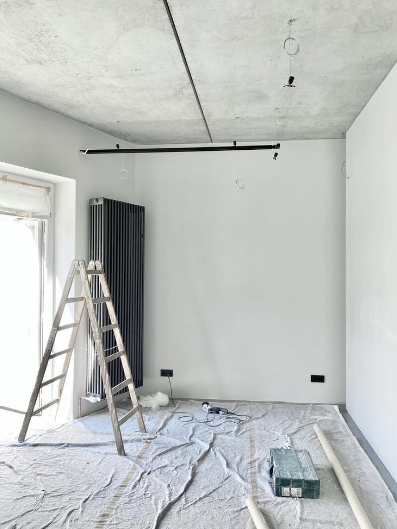 A room undergoing renovation with white walls, a raw concrete ceiling, and a grey drop cloth covering the floor. A wooden ladder leans against the wall next to a dark radiator and a window, while tools and electrical outlets are visible.