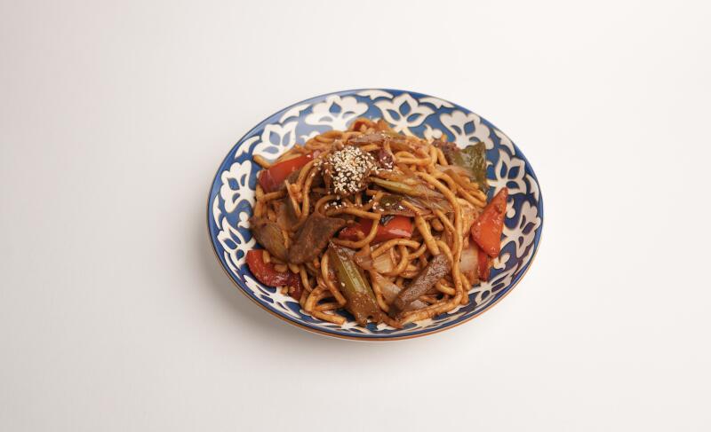 На белой поверхности стоит тарелка лапши с мясом и овощами, посыпанная семенами кунжута. Тарелка имеет сине-белый цветочный узор.