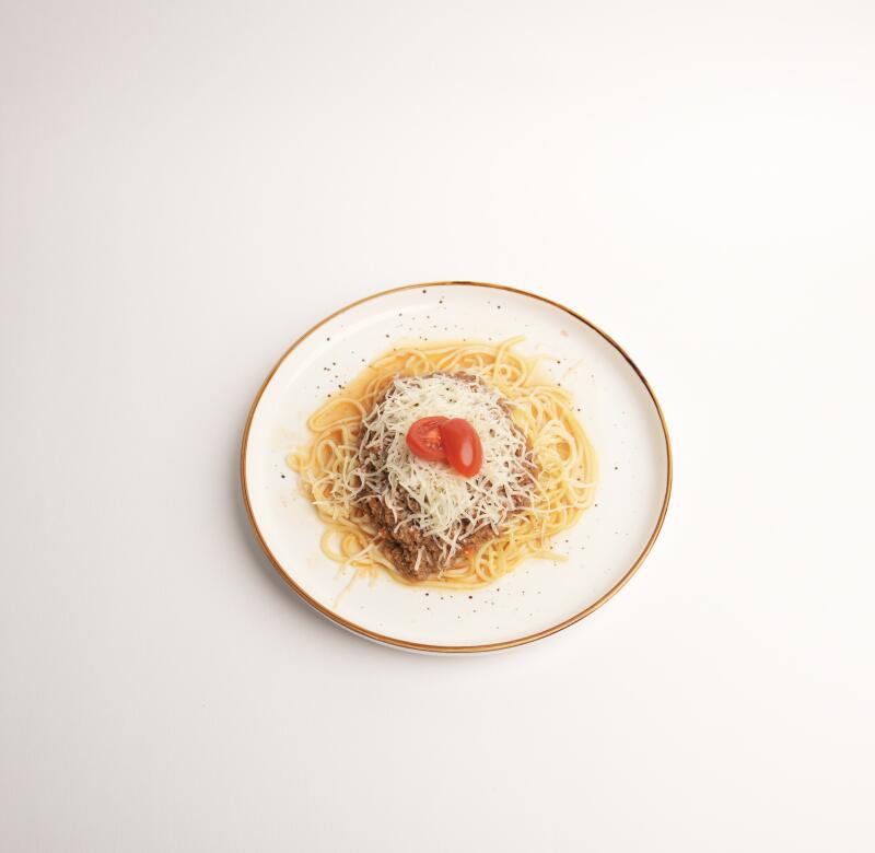 Тарелка спагетти с мясным соусом, тертым сыром и двумя половинками помидора.