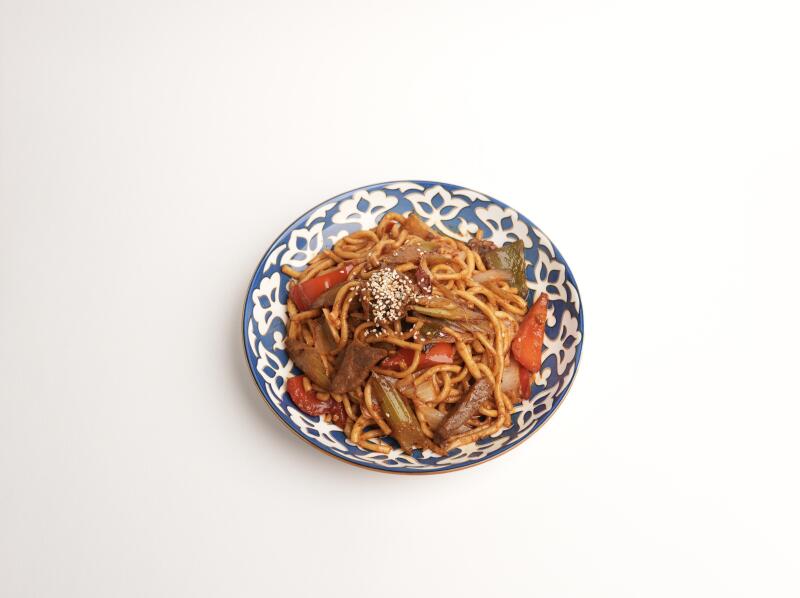 На белом фоне стоит тарелка лапши с овощами и мясом, посыпанная кунжутом. Тарелка имеет сине-белый цветочный узор.