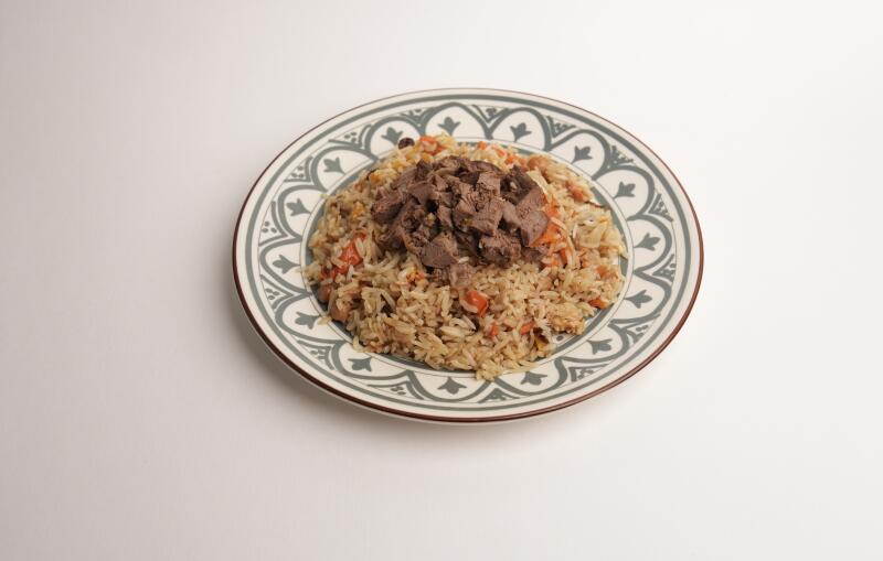 На белой поверхности стоит тарелка плова, сверху которого лежат кусочки мяса. Тарелка украшена декоративным узором в серых и белых тонах.