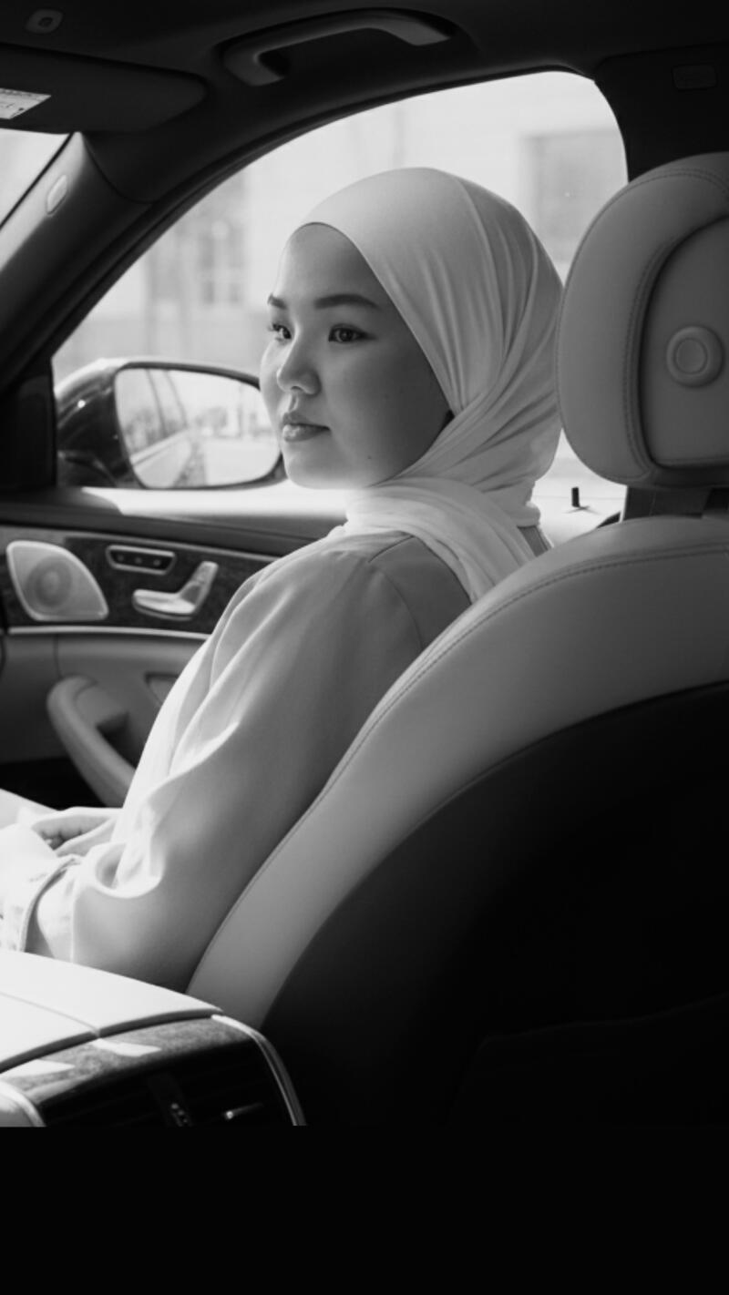 A black and white shot shows a woman in a hijab sitting in a car, looking out the window.
