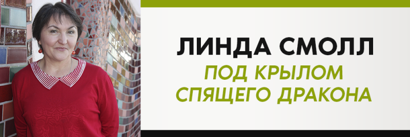 Женщина в красном свитере стоит перед красочной кирпичной стеной, рядом с белой коробкой с текстом "ЛИНДА СМОЛЛ ПОД КРЫЛОМ СПЯЩЕГО ДРАКОНА".