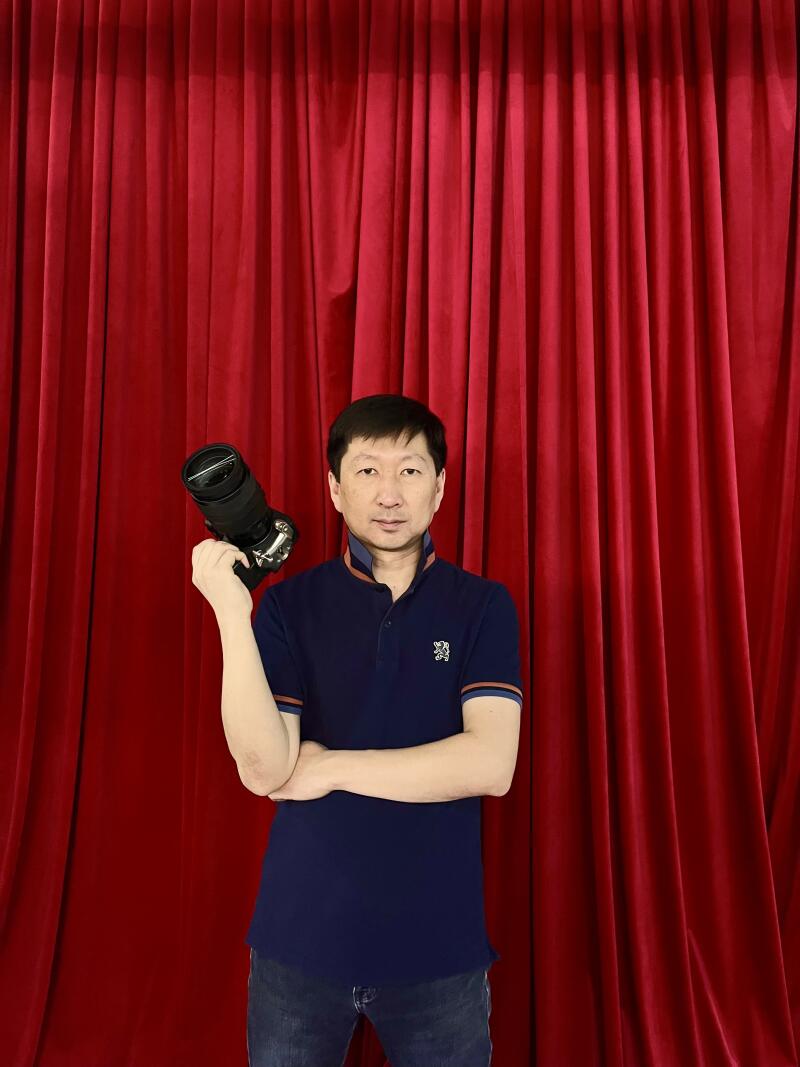 A man in a blue polo shirt holds a camera in front of a red curtain.
