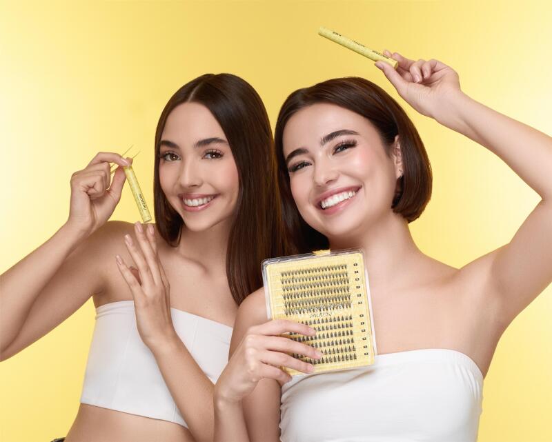 Two smiling women in white tube tops are holding eyelash products against a yellow background. One holds tweezers and a tube, while the other holds a set of individual eyelashes and a tube.