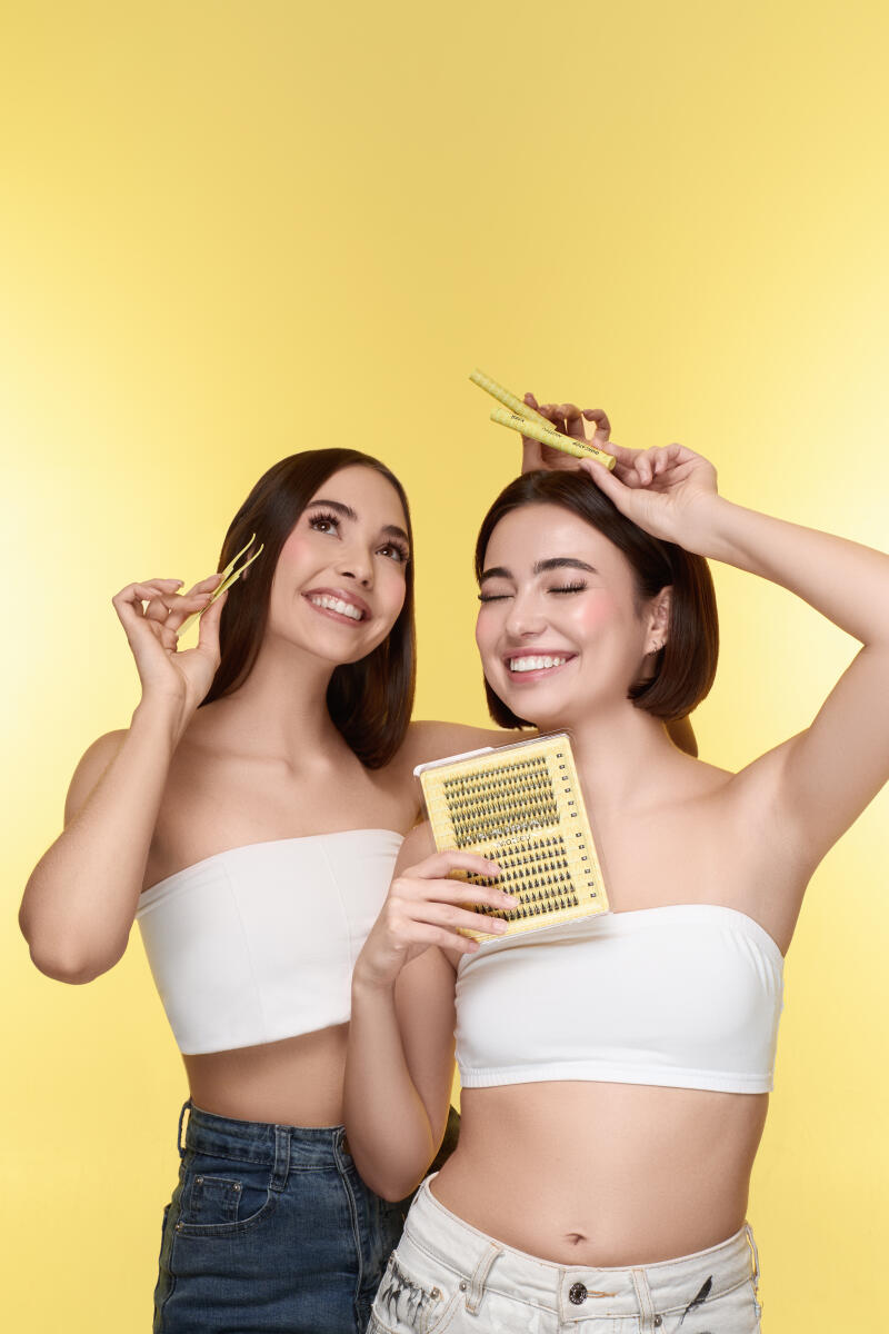 Two smiling women in white tube tops and jeans pose against a yellow background, one holding a lash tray and tweezers, the other holding a lash applicator.