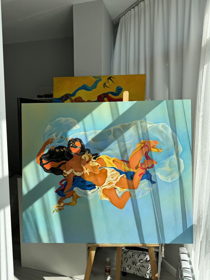 На изображении представлены две картины на мольбертах в комнате с естественным освещением; на передней картине изображены две женские фигуры, окутанные тканью на голубом фоне, а на задней картине изображен желтый фон с синей и красной рекой и птицами.