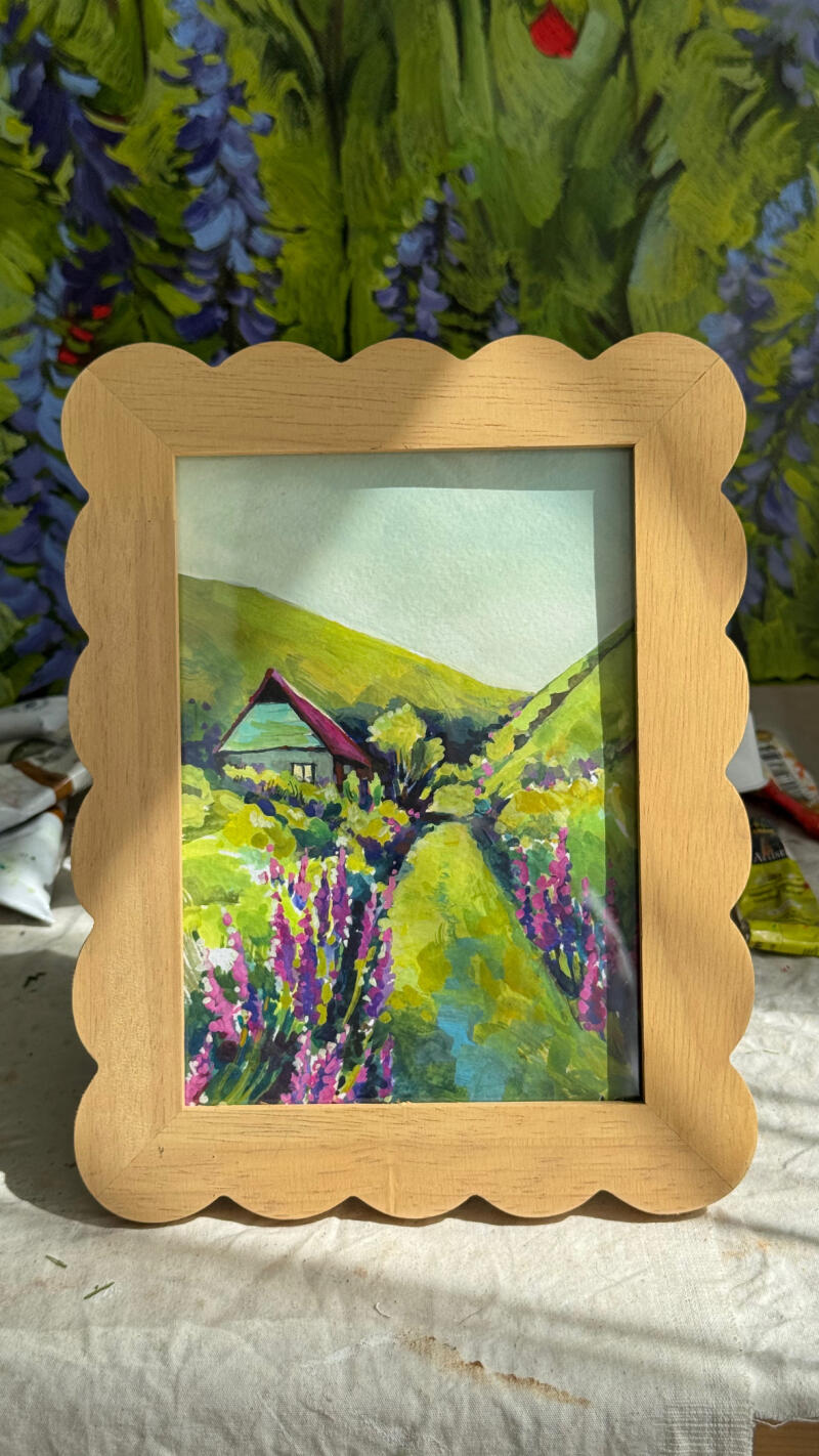 В рамке картина, изображающая пейзаж с маленьким домиком, зелеными холмами и фиолетовыми цветами, обрамляющими тропинку, на фоне цветочной живописи.
