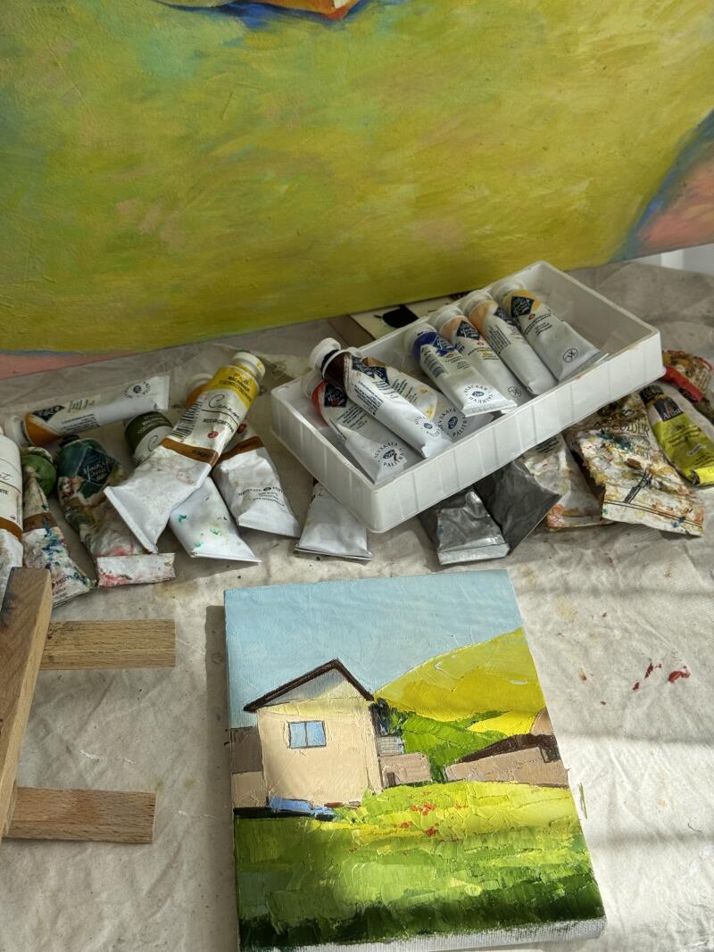 На изображении представлена картина дома на зеленом холме, окруженная тюбиками с краской, некоторые из которых находятся в пластиковом подносе, и деревянными палочками.