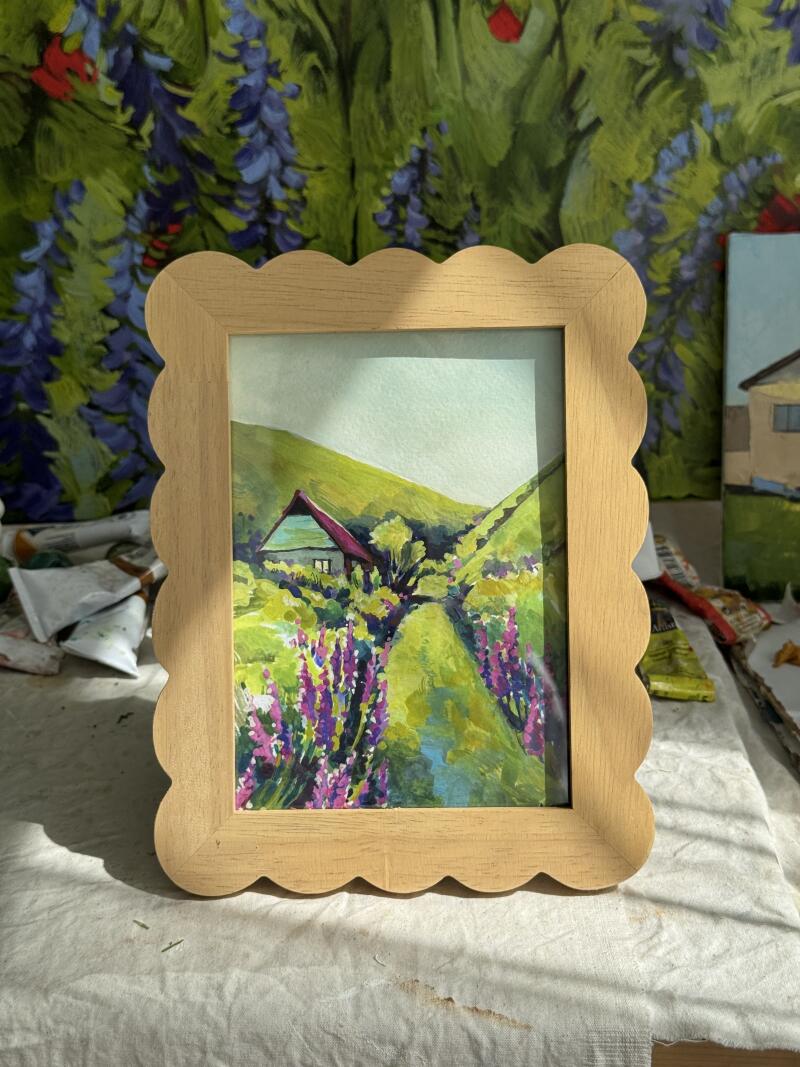 Акварельная картина в рамке с изображением сельского пейзажа с домом, зелеными холмами и фиолетовыми цветами на переднем плане. Рамка светло-коричневая с фестончатым краем.