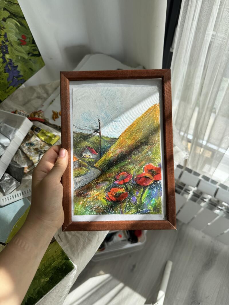Рука держит картину в рамке с изображением пейзажа с красными маками, дорогой, домом и холмом под светло-голубым небом.