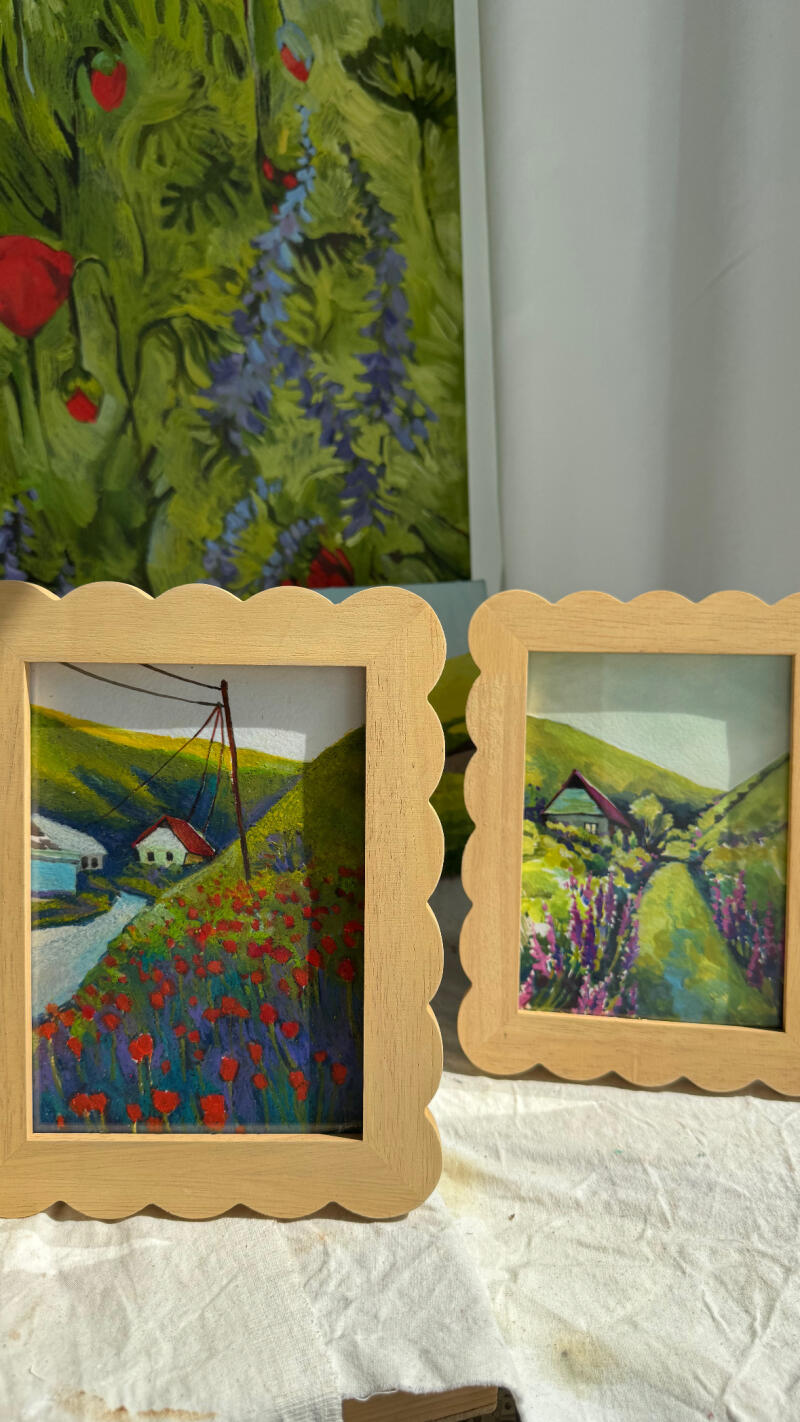 На изображении представлены две маленькие пейзажные картины в рамках и большая цветочная картина на заднем плане. На пейзажных картинах изображены дома и цветы, а на цветочной картине - синие и красные цветы.