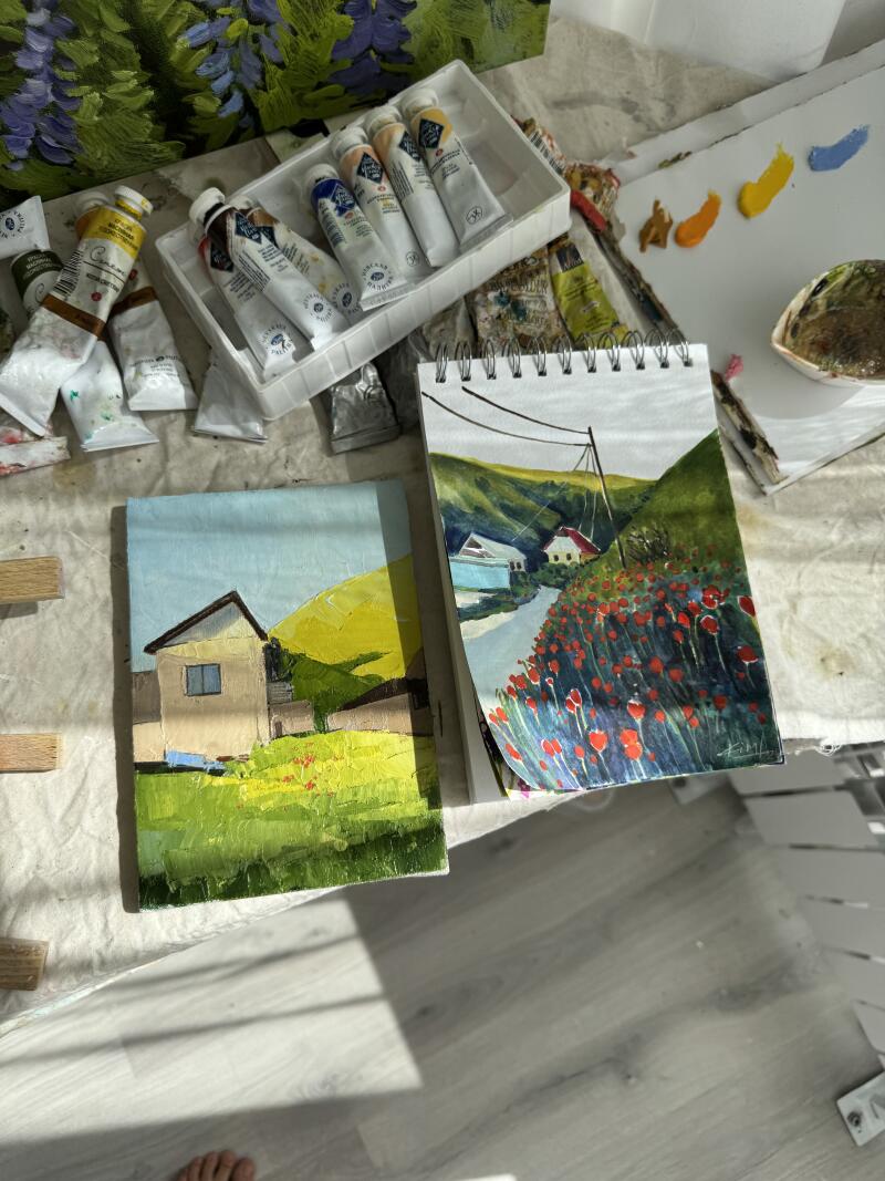 На изображении представлено рабочее место художника с тюбиками краски, двумя картинами с изображением пейзажей с домами и палитрой с мазками краски.
