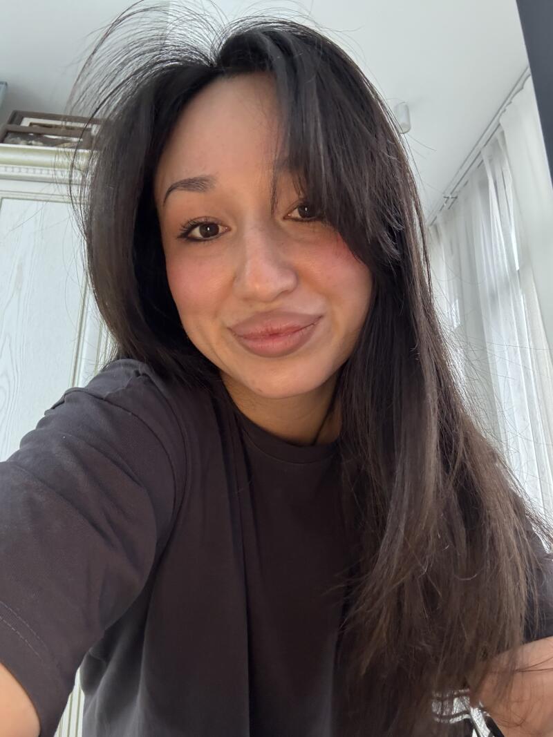 A woman with long dark hair and bangs is wearing a grey t-shirt and smiling at the camera.