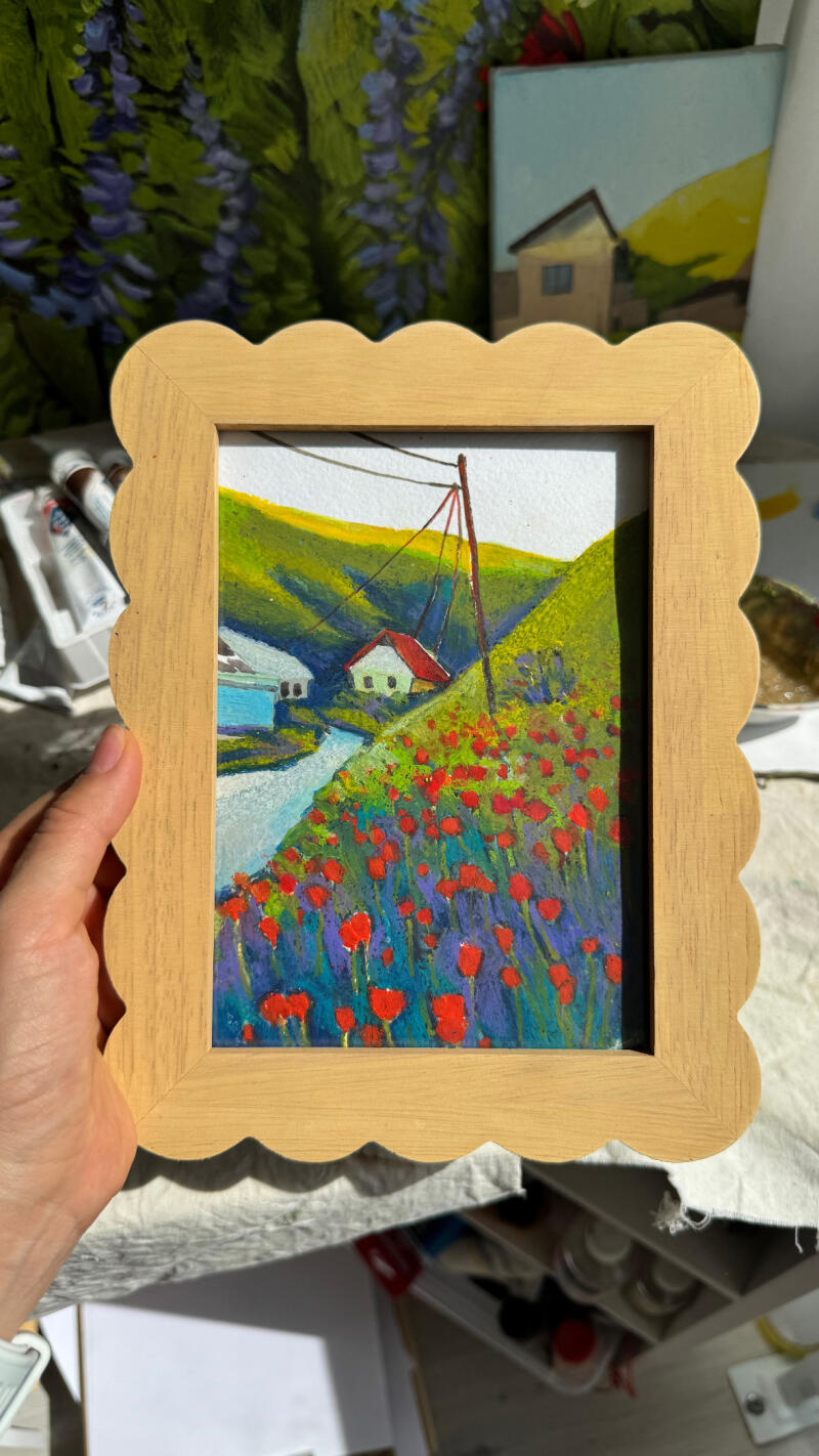 Рука держит картину в рамке с изображением пейзажа с красными цветами, извилистой рекой и домами под зелеными холмами и линией электропередач.