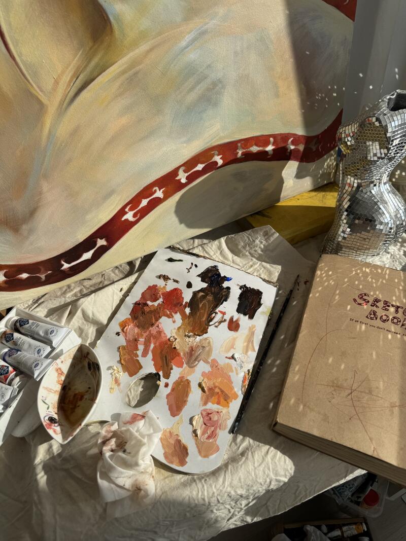 На изображении показано рабочее место художника с холстом, на котором нарисовано изображение, палитрой с различными оттенками коричневой и оранжевой краски, тюбиком краски, альбомом для эскизов и туловищем в виде диско-шара.