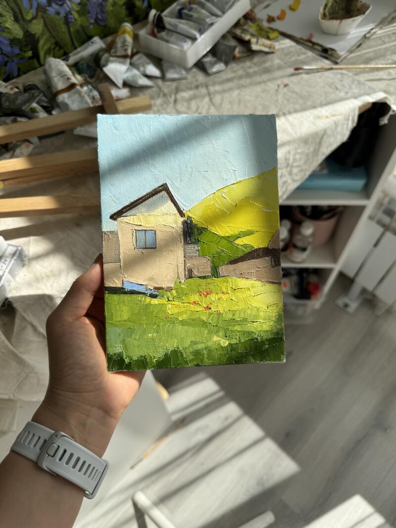 Человек держит пейзажную картину с изображением дома под светло-голубым небом рядом с желтым холмом, с зеленой травой на переднем плане.