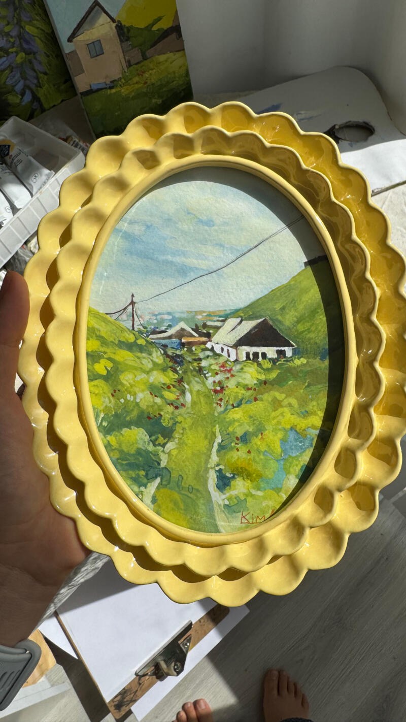 Картина с сельским пейзажем, изображающая дома и зеленые поля, находится в желтой декоративной рамке, на картине видна подпись "Kim".