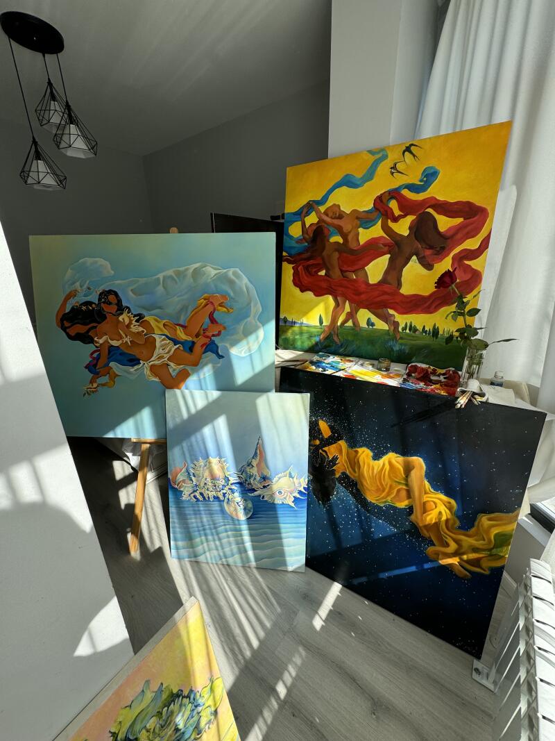 На изображении представлена коллекция ярких картин, прислоненных к стене, демонстрирующих различные темы, включая фигуры в движении с развевающимися тканями, женщину в желтом платье среди звезд и морские ракушки в подводной обстановке.
