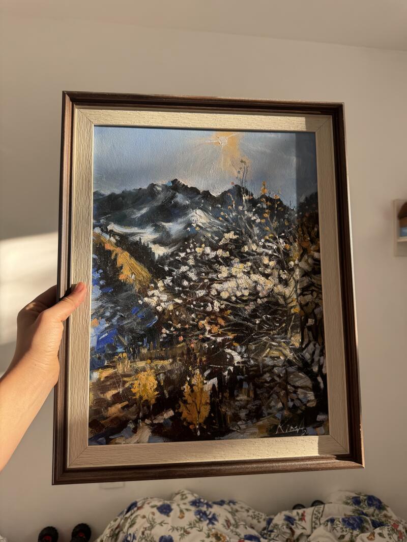 В руках держат обрамлённую масляную картину с горным пейзажем, на которой изображены заснеженные горы, деревья с белыми цветами и осенняя листва.