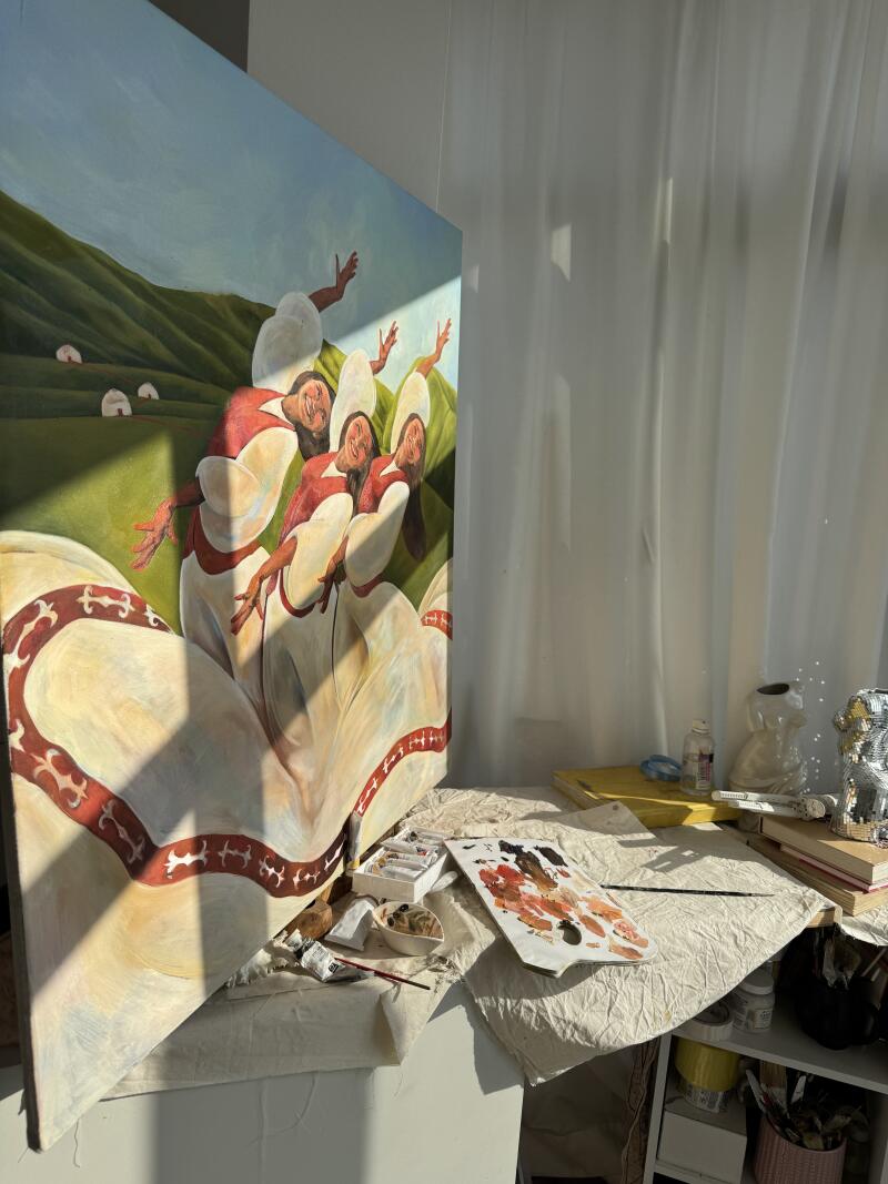 На изображении картина с изображением четырех женщин в белых платьях с красными деталями на зеленом склоне холма, а рядом на столе лежат художественные принадлежности.