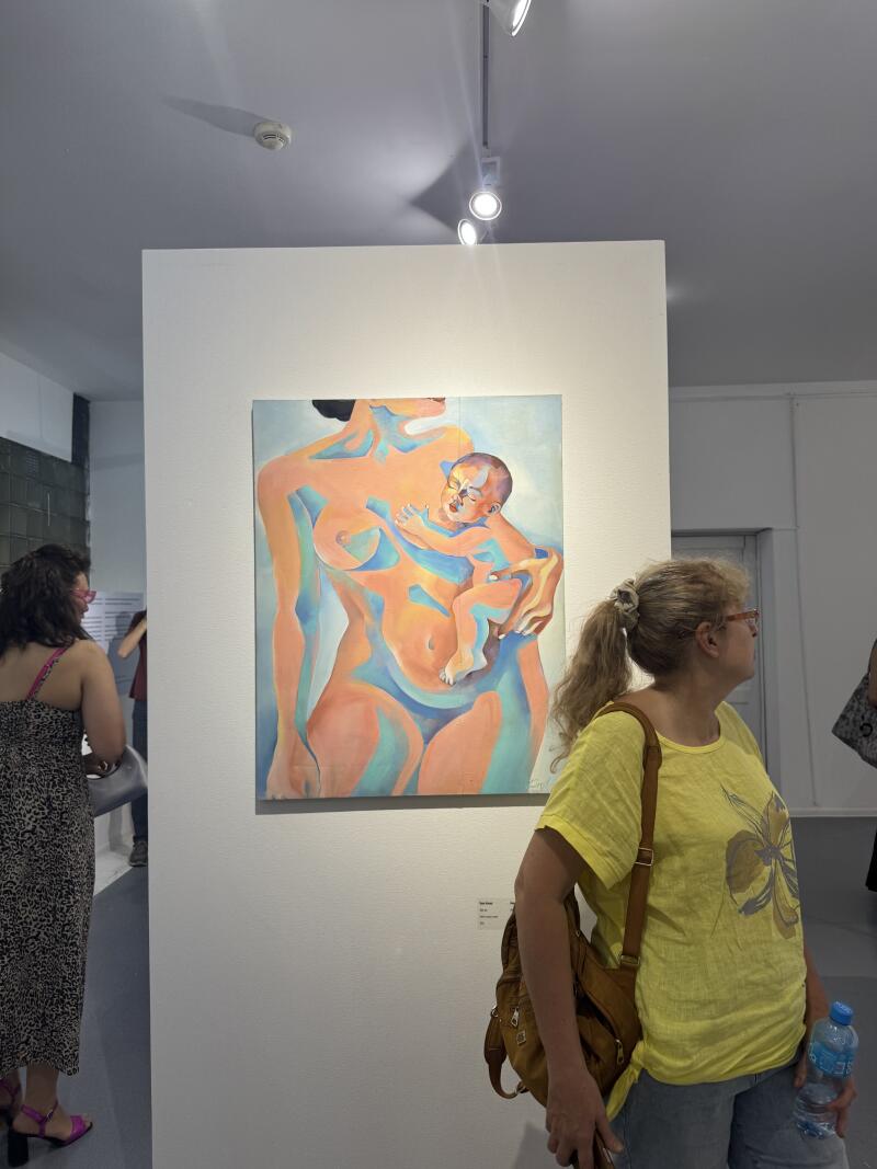 Картина обнаженной женщины, держащей ребенка, висит в галерее, а две женщины стоят рядом и рассматривают произведение искусства.