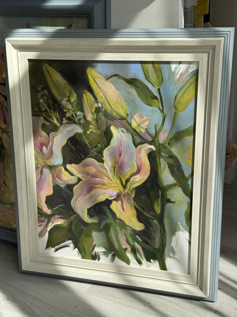 На картине в рамке изображены лилии в оттенках желтого, розового и зеленого на голубом фоне.