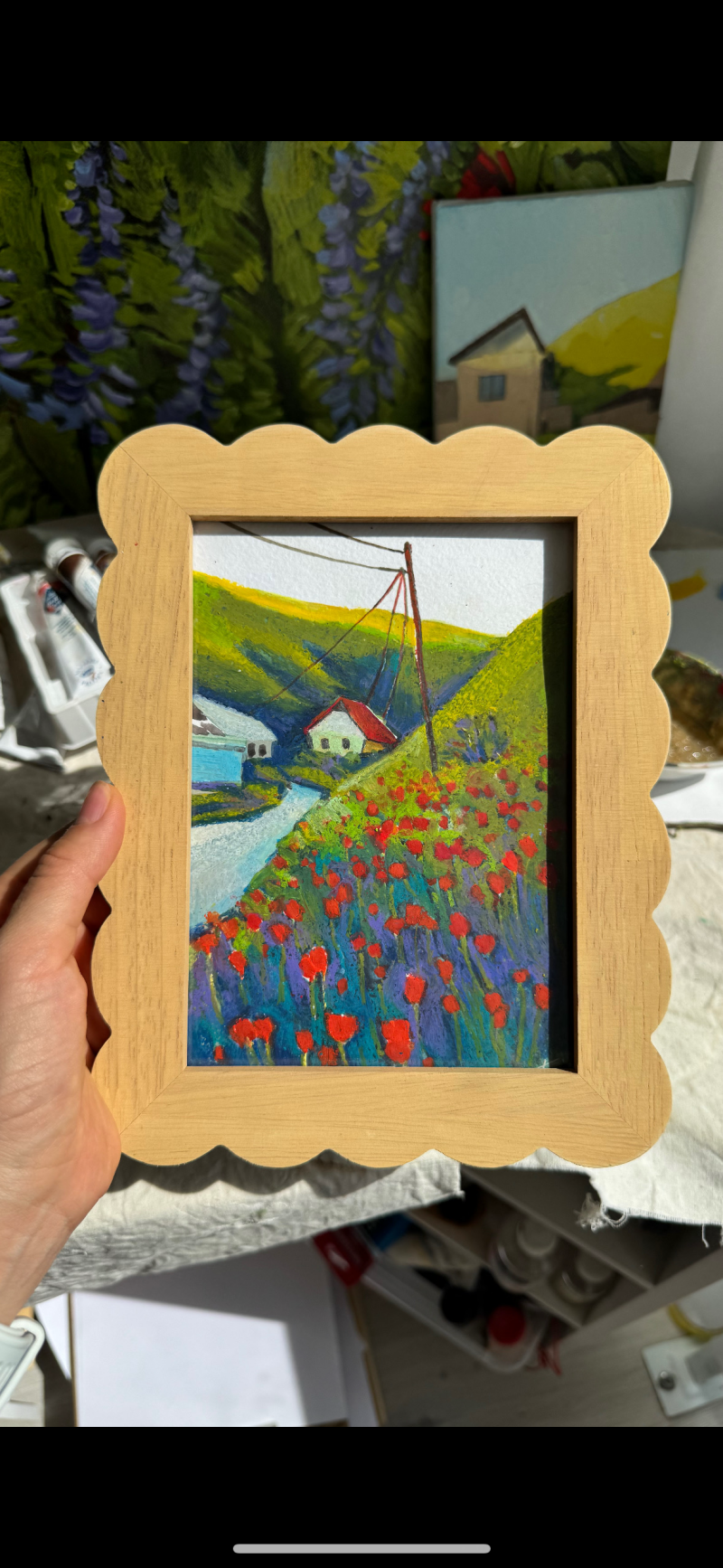Человек держит картину в рамке с изображением сельского пейзажа с красными цветами на переднем плане, извилистым ручьем, домами и зелеными холмами на заднем плане, под электрическим столбом.
