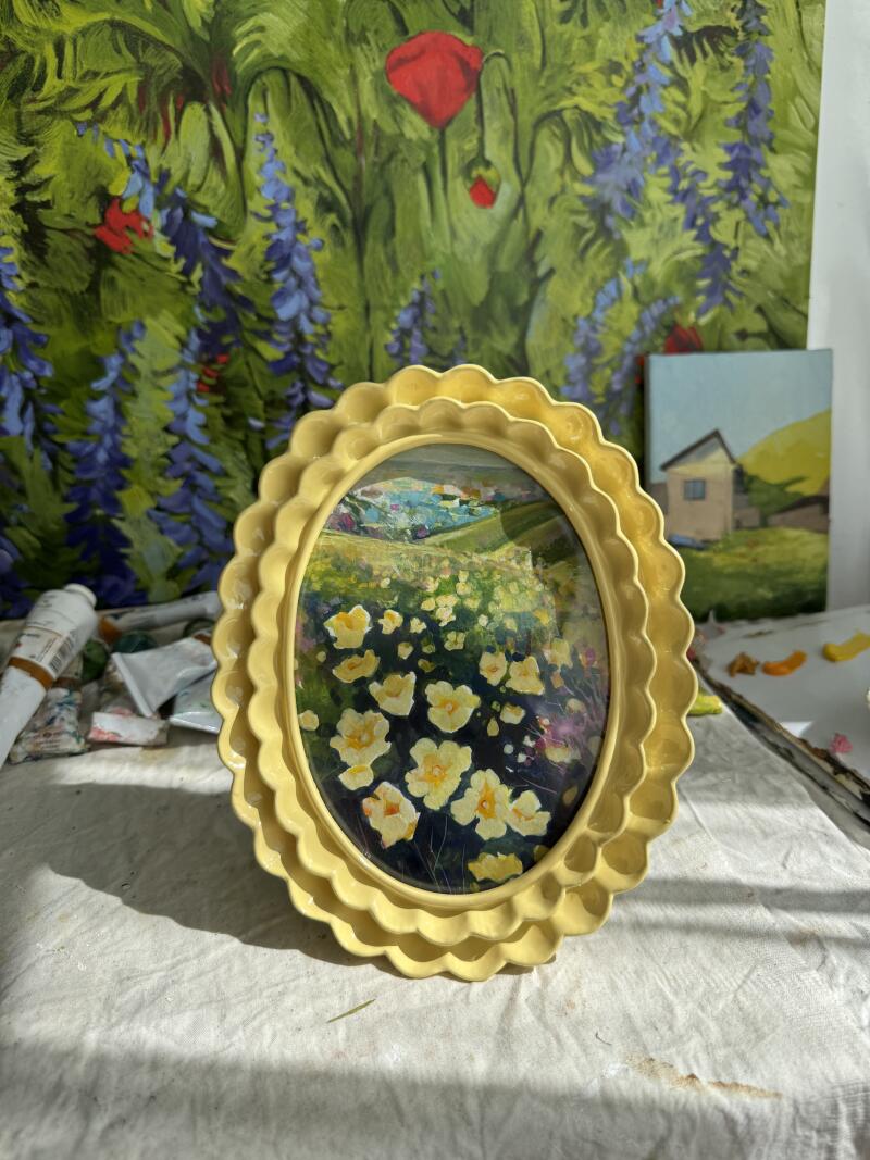 На изображении картина в жёлтой рамке с изображением жёлтых цветов на переднем плане и пейзажа на заднем плане, а также художественные принадлежности и другие картины на заднем плане.