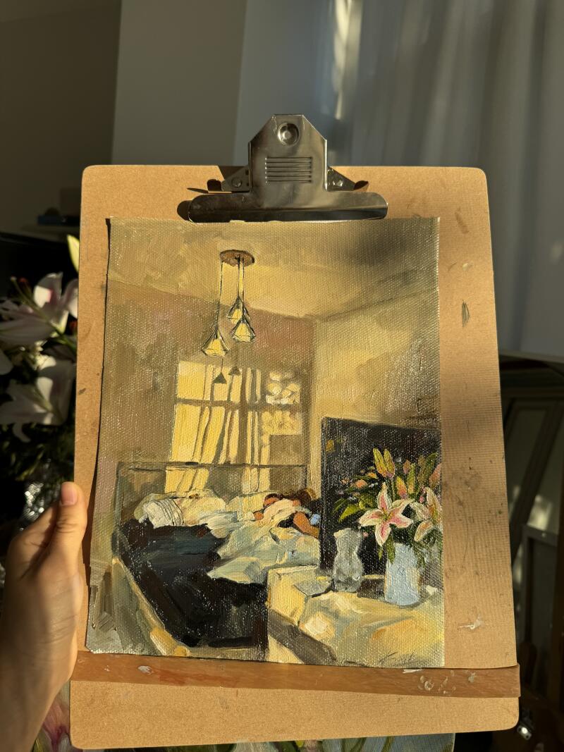 Картина с изображением спальни закреплена на планшете, на ней изображена кровать с постельным бельем, окно с проникающим сквозь него светом, прикроватный столик с цветами и подвесной светильник.